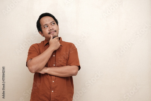 Southeast Asian man in a brown shirt standing with hand on chin, looking up thoughtfully as if pondering or considering an idea.
