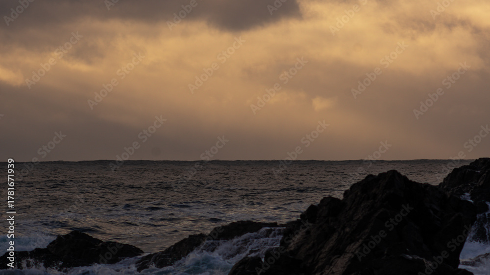 Obraz premium Rough ocean waters surge against jagged coastal rocks beneath an intensely cloudy, ochre-tinted sky at dawn or dusk.
