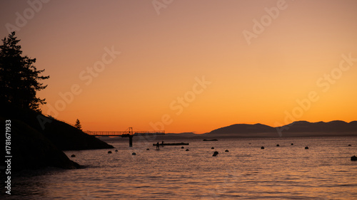 A Tranquil Sunset Paints the Coastal Sky with Warm Orange Hues, Silhouetting a Pier, Paddleboarders, and Distant Mountains