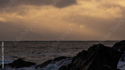 Rough ocean waters surge against jagged coastal rocks beneath an intensely cloudy, ochre-tinted sky at dawn or dusk.