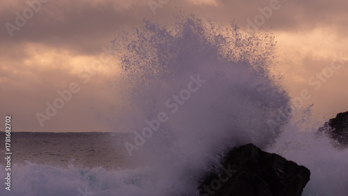Powerful Ocean Wave Exploding Against Dark Jagged Coastal Rocks Under a Warmly Lit Cloudy Sky at Twilight