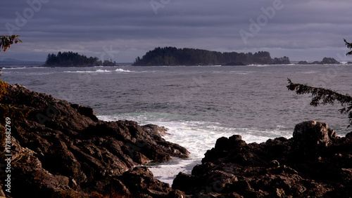 Wild Pacific Ocean Waves Crash Against Rugged Dark Coastal Rocks, Framing Distant Tree-Covered Islands Under a Moody, Overcast Sky.