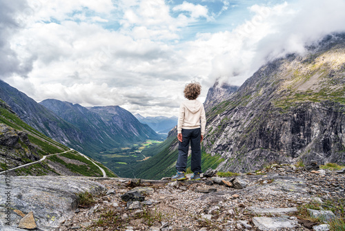 A young boy stands at the edge of a deep valley, gazing at the stunning landscape of Trollstigen mountain pass in Norway, surrounded by majestic mountains. Rear view
