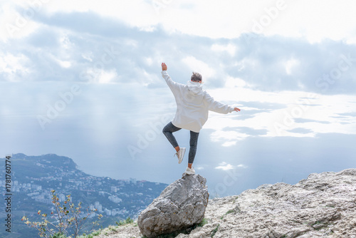 A man stands on one leg on a rock high in the mountains. A young woman in a white hoodie and black jeans looks down at the city and the sea. A magnificent view of beautiful nature. 