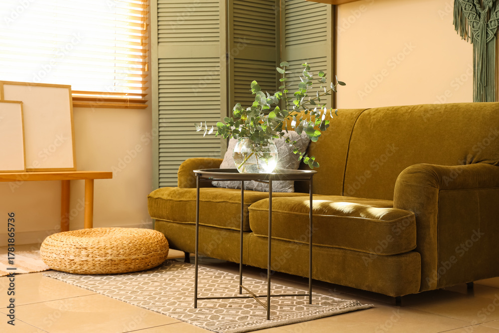 Obraz premium Glass vase with fresh eucalyptus branches on table near green sofa and folding screen in living room