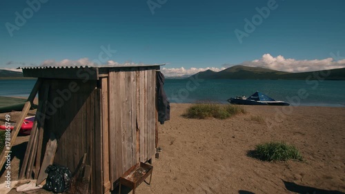 Rough-plank shed next to kayaks and inflatable raft on sandy shore of sea bay surrounded by green mountain ranges. Travel and tourism concept