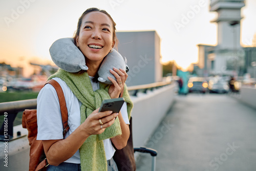 Quadro em tela Young woman traveler smiling, holding smartphone and wearing a neck pillow while