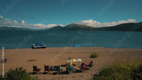 Three young people sit on folding chairs on sandy beach near table with thermoses and containers of food and admire sea bay with ridges of mountains. Back view