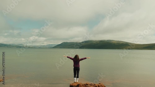 Young woman stands at water's edge of sea bay with arms spread out to sides and enjoys freedom and majestic beauty of mountains on horizon. Back view
