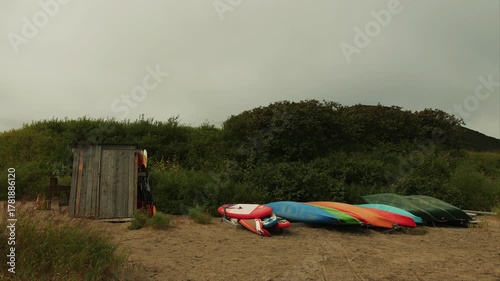 Inflatable boats, kayaks, and air mattresses dried upside down on shore next to small shed made of rough planks. Wind sways grass and bushes on green hills