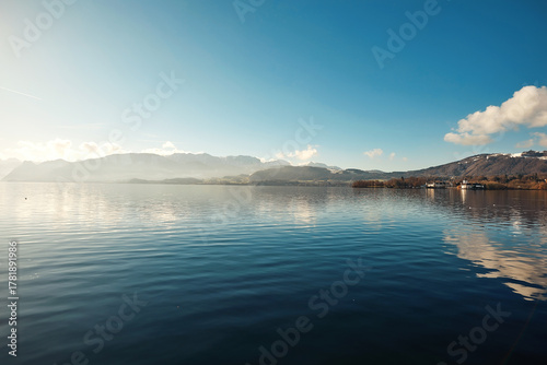 Serene lake landscape with calm waters reflecting mountains and clouds under a clear blue sky, showcasing natural beauty and tranquility
