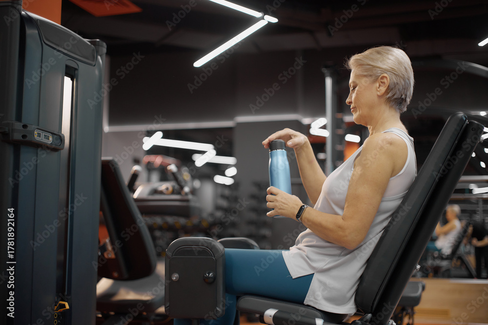 Fototapeta premium Senior Caucasian woman exercising on leg press machine in gym, holding water bottle and taking break during workout, short gray hair visible, focused on fitness routine
