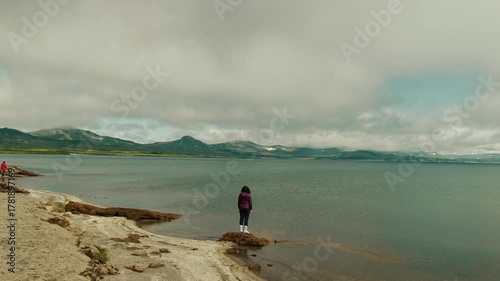 Young woman stands at water's edge of sea bay with arms spread out to sides and enjoys freedom and majestic beauty of mountains on horizon.