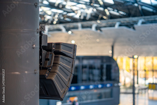 Public address speaker array mounted on metal column inside modern transportation building, part of passenger voice announcement and audio communication system