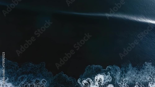 Aerial view of dark blue ocean with white foamy waves washing onto a black sand shore.
