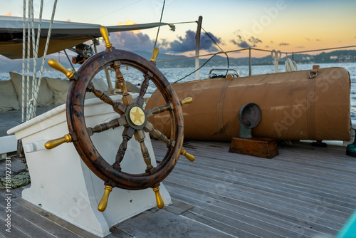 An unmanned wooden yacht wheel with gold handles glows in the Caribbean sunset, the island beyond bathed in warm light and tranquillity