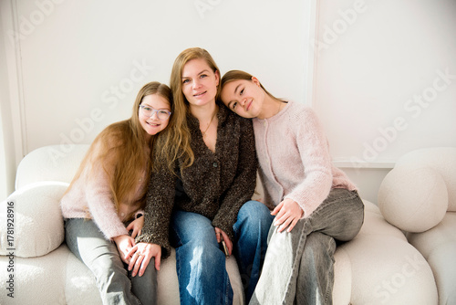 beautiful blonde smiling mother and daughter with long hair sitting on sofa in white studio