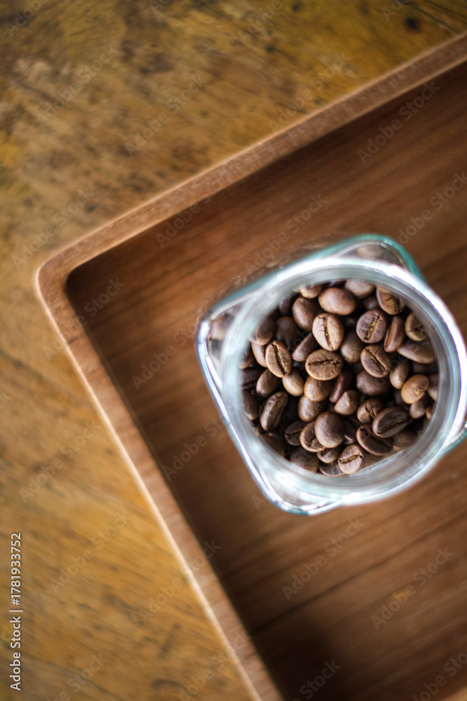 Fototapeta premium Roasted coffee beans spilling from a glass jar onto a wooden surface under warm natural light. 