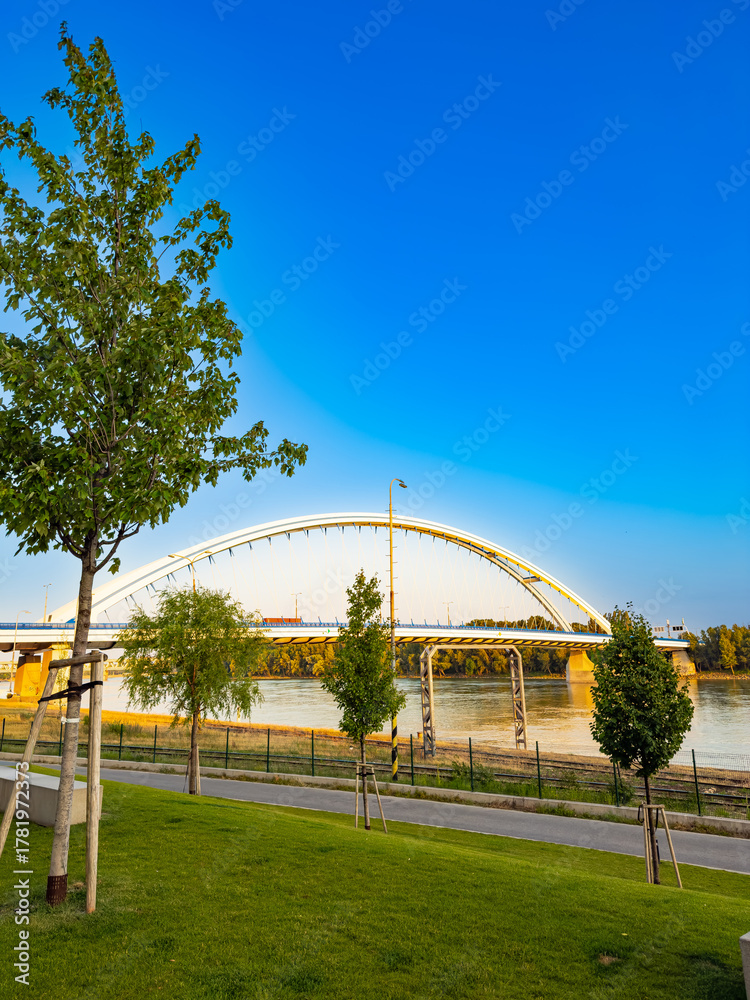 Naklejka premium White arch bridge over Danube River in Bratislava Slovakia. Scenic modern bridge crossing the Danube with green park foreground and blue sky