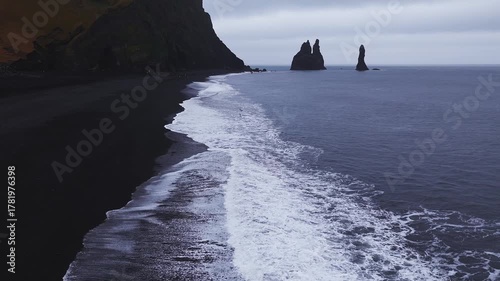 Aerial view shows Reynisdrangar sea stacks at Reynisfjara near Vik, Iceland. Foamy waves wash black sand by basalt cliffs, tiny visitors add scale under overcast light.