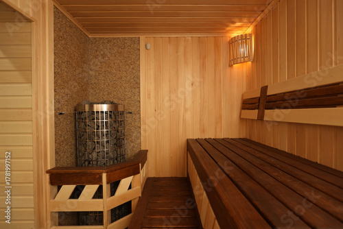 Warm cozy sauna interior with wooden walls, ceiling and floor, featuring a stove with hot stones in the corner. Soft and dim lighting creates a relaxing and intimate atmosphere.