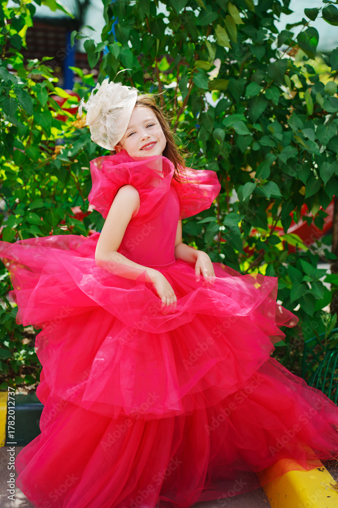 Fototapeta premium A girl in a lush pink dress and hat near the green bushes