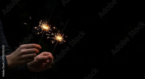 Hands Holding Two Sparklers Glowing Brightly Against a Deep Black Background