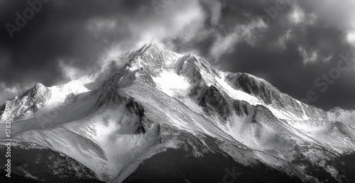 Snow-covered mountain peaks under dramatic cloudy sky with dark forest at base creating a moody atmosphere