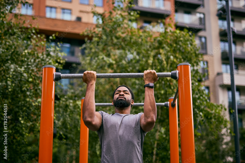 Fototapeta premium Man Doing Pull-Ups At Outdoor Gym In City Park, Focused On Training And Fitness