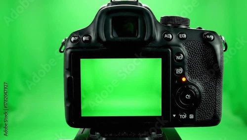 A black DSLR camera with a green screen display faces forward against a vibrant green backdrop, illuminated by soft, even lighting.

