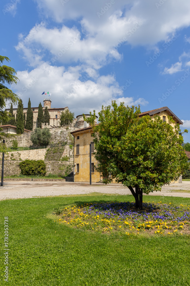 Obraz premium View of Brescia Castle from Piazzale della Locomotiva, framed by a green lawn and summer light highlighting the medieval fortress