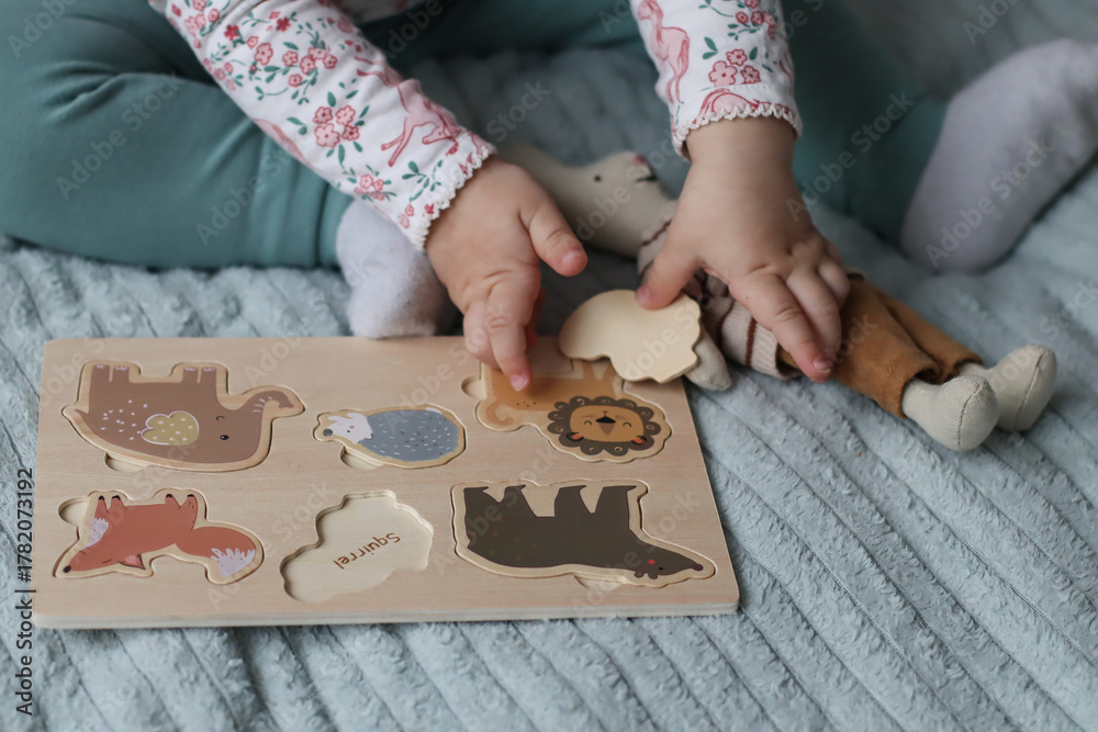 Obraz premium Close-up of child’s hands assembling a wooden animal puzzle on a soft surface indoors. Educational toy for early learning, fine motor skills, and creative play in a cozy home environment.