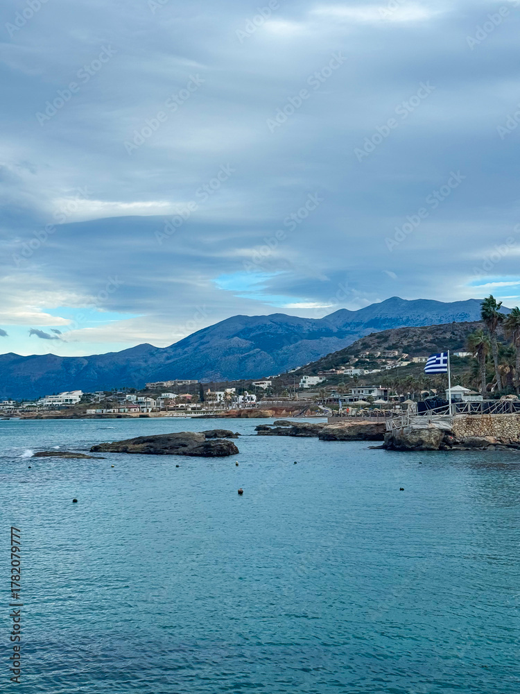 Naklejka premium Scenic coastal view showcasing a tranquil shoreline and mountainous backdrop under a cloudy sky during a calm afternoon in Greece