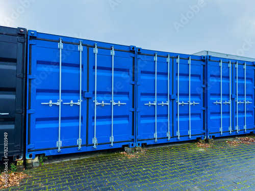 Bright blue shipping containers lined up against a grey sky in a storage yard on an overcast day