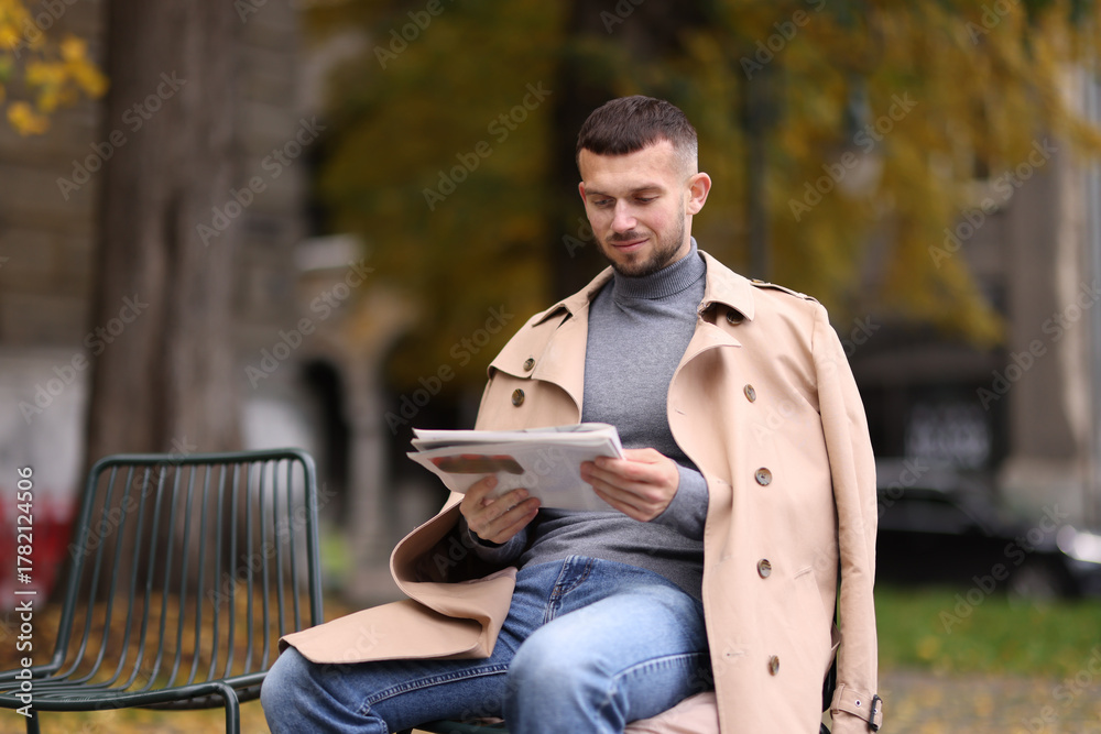 Obraz premium Young man reading newspaper on chair outdoors