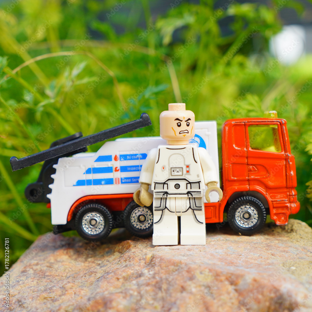 Obraz premium Lego figure stands next to a toy tow truck on a rock with green foliage