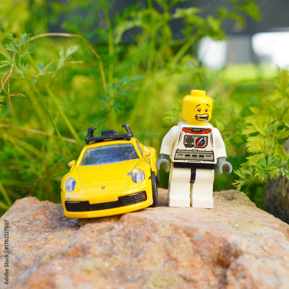 Obraz premium Lego astronaut and yellow toy car on a rock with green foliage background