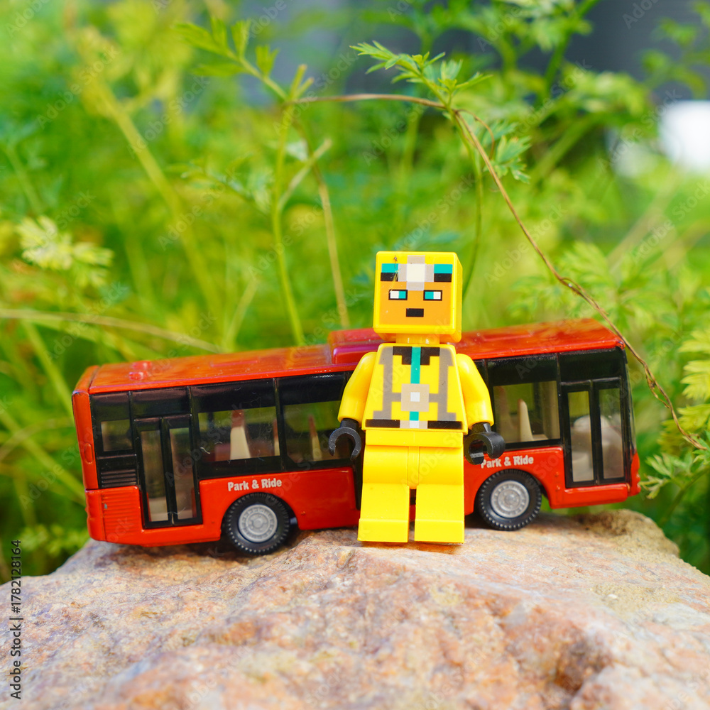 Naklejka premium Lego figure stands in front of a red toy bus on a rock with green foliage