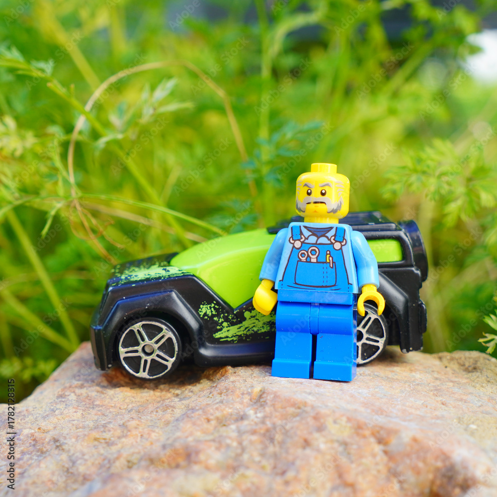Naklejka premium Lego mechanic figure stands next to a toy car on a rock in a garden
