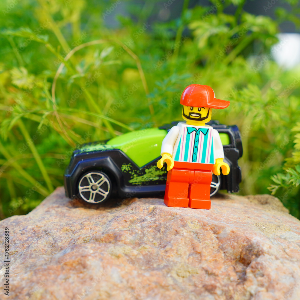 Naklejka premium Lego minifigure stands next to a toy car on a rock in a natural setting