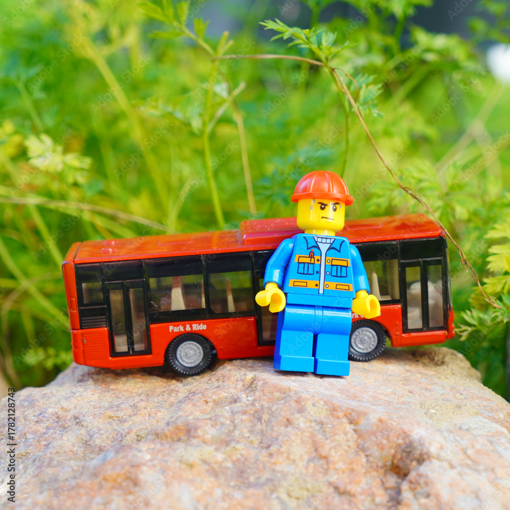 Naklejka premium Lego figure with a red bus on a rock in front of green foliage