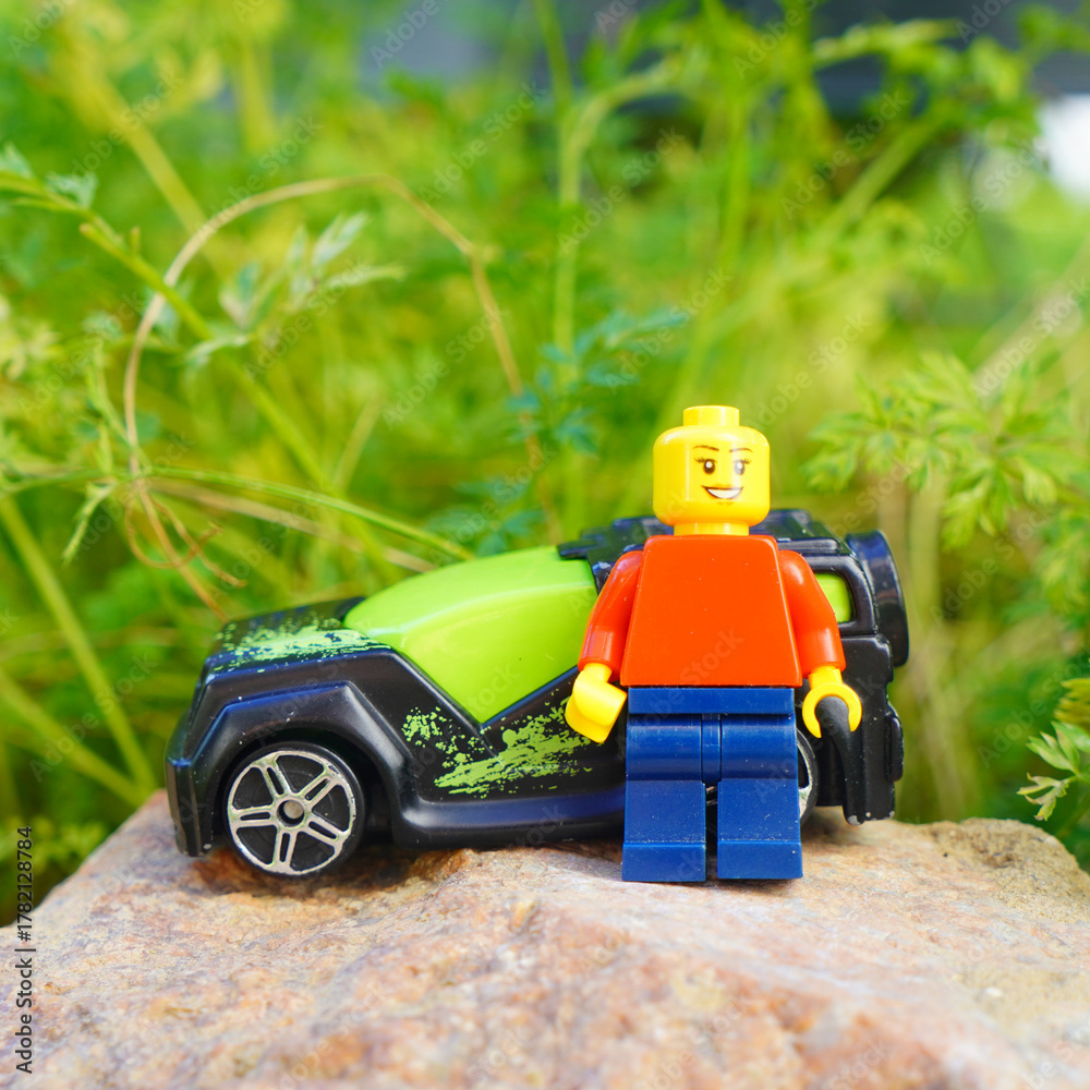 Naklejka premium Lego minifigure stands beside a toy car on a rock with a green leafy background