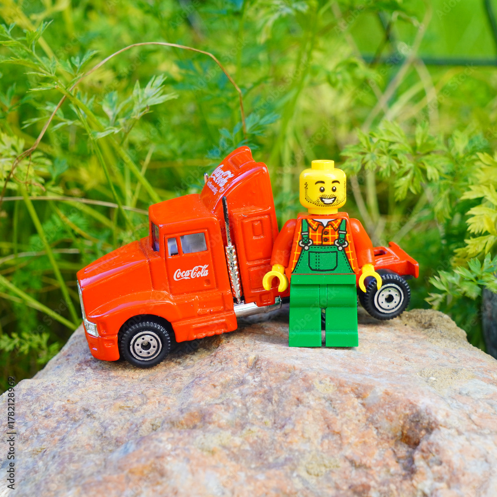 Naklejka premium Lego figure with Coca-Cola truck on a rock with green foliage background