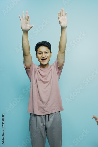 Cheerful young man raising his hands in celebration against a vibrant teal background, radiating positivity and youthful energy, perfect for lifestyle ads