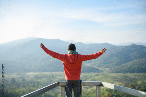 Man in an red hoodie stands on a viewpoint with arms outstretched, facing a vast, misty mountain landscape, symbolizing freedom, success, nature, and adventure.