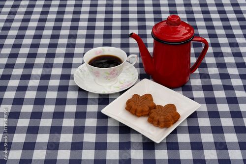 Top view of a red coffee pot, a coffee cup, and a plate with two maple leaf-shaped Wagashi pastries on a navy blue and white plaid tablecloth. Ideal for autumn snack and Japanese food concept.