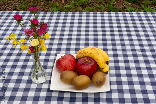 Top view of a platter of fruit (apples, kiwis, bananas) and a small glass vase with a vibrant bouquet on a navy blue and white plaid tablecloth. Ideal for a healthy snack or brunch.