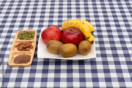 Platter of fruit (apples, kiwis, bananas) and a divided tray with almonds, pumpkin seeds, and flax seeds on a navy blue and white plaid tablecloth. Ideal for healthy snack and diet concepts.