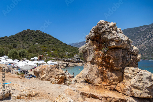 Fototapeta Naklejka Na Ścianę i Meble -  Scenic rocky outcrop near Kaş peninsula with turquoise sea, green hills, İnceboğaz beach with white umbrellas, and forested mountains in haze. Mediterranean resort vibe. Turkey.

