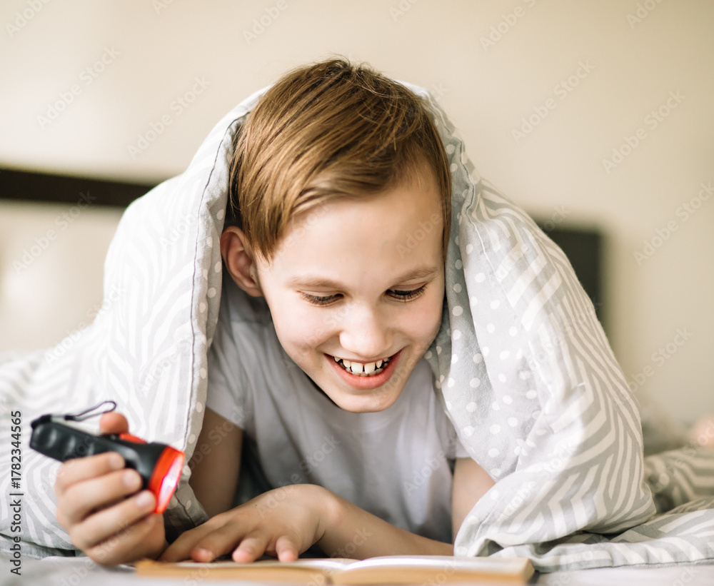 Fototapeta premium Cute kid boy teenager reading a book under the covers with flashlight 